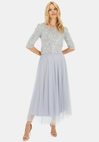 Srebrna bluzka z cekinami, mająca trzy czwarte rękawy, połączona z jasnoszarym tiulowym spódnicą, charakteryzującą się zwiewną sylwetką. Całość dopełniają cieliste szpilki.