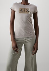 Camiseta ajustada de color beige claro con un parche rectangular texturizado con "A X" en negro en la parte delantera. Mangas cortas y bordes sin costuras.