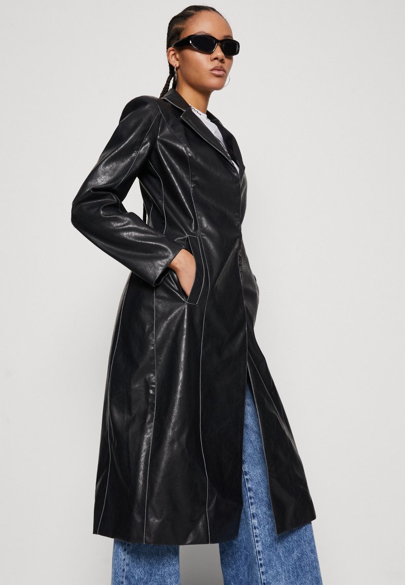 Abrigo largo de cuero negro con un diseño a medida, que presenta costuras blancas, cuello con muesca y bolsillos laterales, llevado sobre pantalones de mezclilla azul claro.