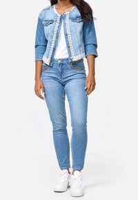 Chaqueta de denim con mangas tres cuartos, lavado azul claro, detalles de encaje, combinada con jeans ajustados azul claro y zapatillas blancas.