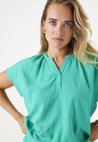 Türkiser kurzärmeliger Oberteil mit V-Ausschnitt, strukturiertem Stoff und sanften Falten an den Schultern. Leicht lockere Passform.