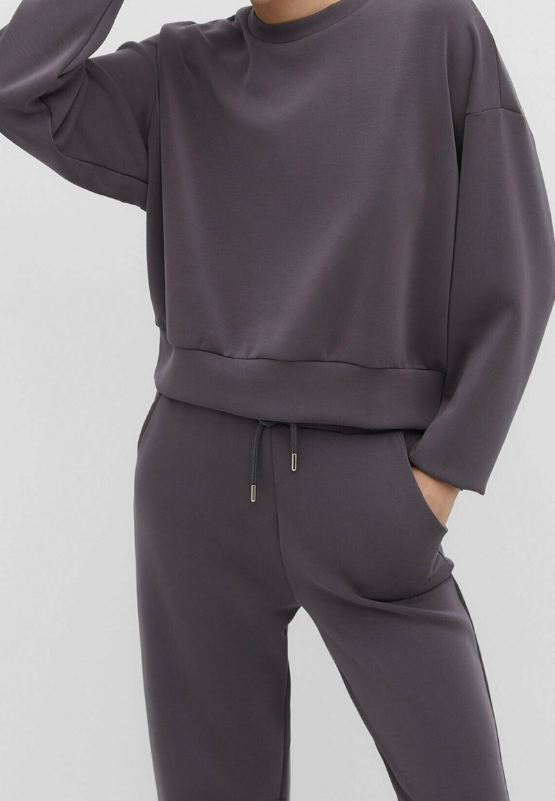 Personne portant un sweat-shirt gris foncé à manches longues et un pantalon à cordon assorti, une main dans la poche, debout devant un fond uni.