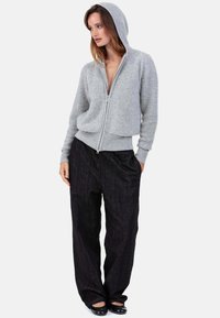 Sweat à capuche gris à fermeture éclair avec une finition texturée et une capuche, associé à un pantalon noir ample avec des rayures discrètes et des poches latérales. Chaussures plates visibles.