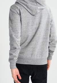 Sudadera gris con acabado texturizado, capucha con cordón y puños de canalé. Combinada con pantalones negros, vista desde la parte posterior.