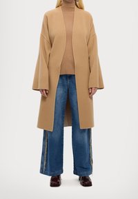 Manteau oversize en mélange de laine beige avec des manches larges, porté sur un col roulé beige, associé à un jean en denim bleu évasé avec des détails argentés sur les côtés.