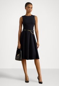 Melna kleita bez piedurknēm ar zvilni svārku, akcentētu ar melnu jostu ar zelta ķēdi. Modele tur melnu rokassomu un valkā melnas augstpapēžu kurpes.