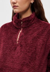 Bordowy polar z wysokim kołnierzem i półzamkiem; cechuje się teksturowaną, miękką powierzchnią oraz kontrastującym jasnobeżowym detalem zamka.