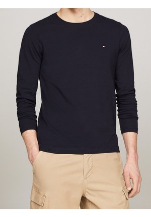 Homme portant une chemise ajustée bleu marine à manches longues et un pantalon cargo beige, les mains dans les poches, debout devant un fond neutre.