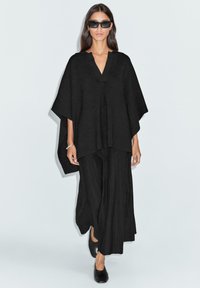 Femme portant un poncho noir surdimensionn�é, un pantalon noir à jambes larges, des chaussures noires, des lunettes de soleil foncées et un bracelet argenté, marchant sur un fond blanc uni.