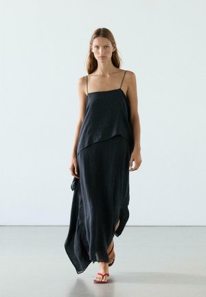 Kobieta idąca do przodu, ubrana w czarną, długą sukienkę na ramiączkach i czerwone sandały, na jasnym, jednolitym tle.