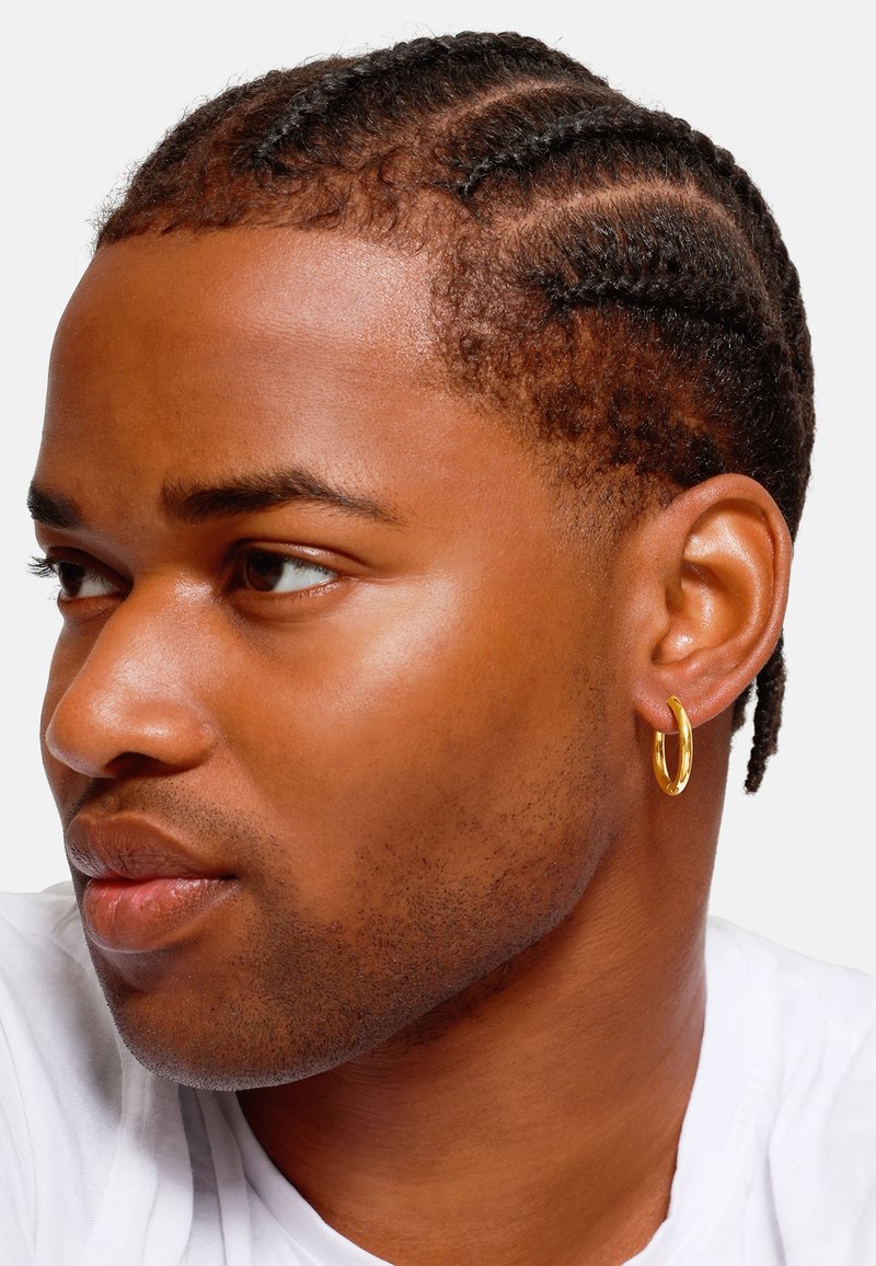 Boucle d'oreille en or sur une personne avec des cheveux tressés, peau claire et t-shirt neutre ; en mettant l'accent sur le design circulaire et brillant de la boucle.
