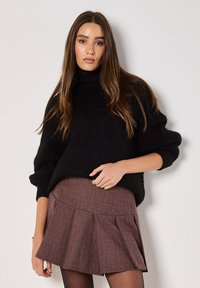Schwarzer gestrickter Pullover mit hohem Kragen, kombiniert mit einem karierten Skaterrock in Burgunder- und Brauntönen; das Model steht mit einer Hand an der Hüfte.