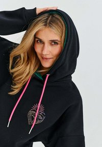 Černá mikina s růžovými šňůrkami, zelenou podšívkou a srdcovým designem se vlnami a sluncem v pastelových barvách. Měkká tkanina, volný střih.