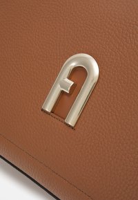 Hnědá kožená taška s texturovaným povrchem a výrazným zlatým kovovým logem. Viditelné švy se nacházejí podél okrajů.