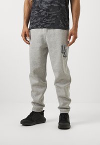 Grå bomulds sweatpants med tætsiddende manchetter, med et stort "UA" logo i sort på venstre lår og kombineret med sorte sportssko.