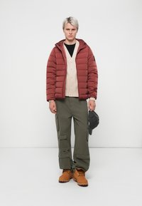 Puffjacke in rostrot, gestepptes Design, mit Kapuze, über einem hellbeigen Cardigan. Olivgrüne Cargo-Hosen, braune Stiefel, graue Mütze haltend.