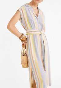 Abito leggero a righe in blu, giallo e beige con colletto, bottoni e cinta in vita. Abbinato a una borsa intrecciata e accessori.