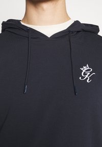 Námořnická mikina s jemnou texturou, s bílým vyšívaným logem a designem koruny, a nastavitelnými šňůrkami na kapuci.