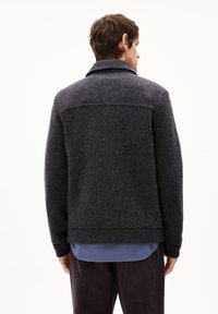 Dunkelgrau gestrickte Jacke mit hohem Kragen, gerippten Bündchen und glatter Textur. Über einem hellblauen Hemd getragen, was einen gestuften Look zeigt.