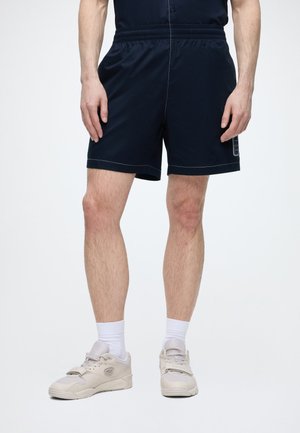 Homme portant un short noir, des chaussettes blanches et des baskets beiges, debout devant un fond clair uni.