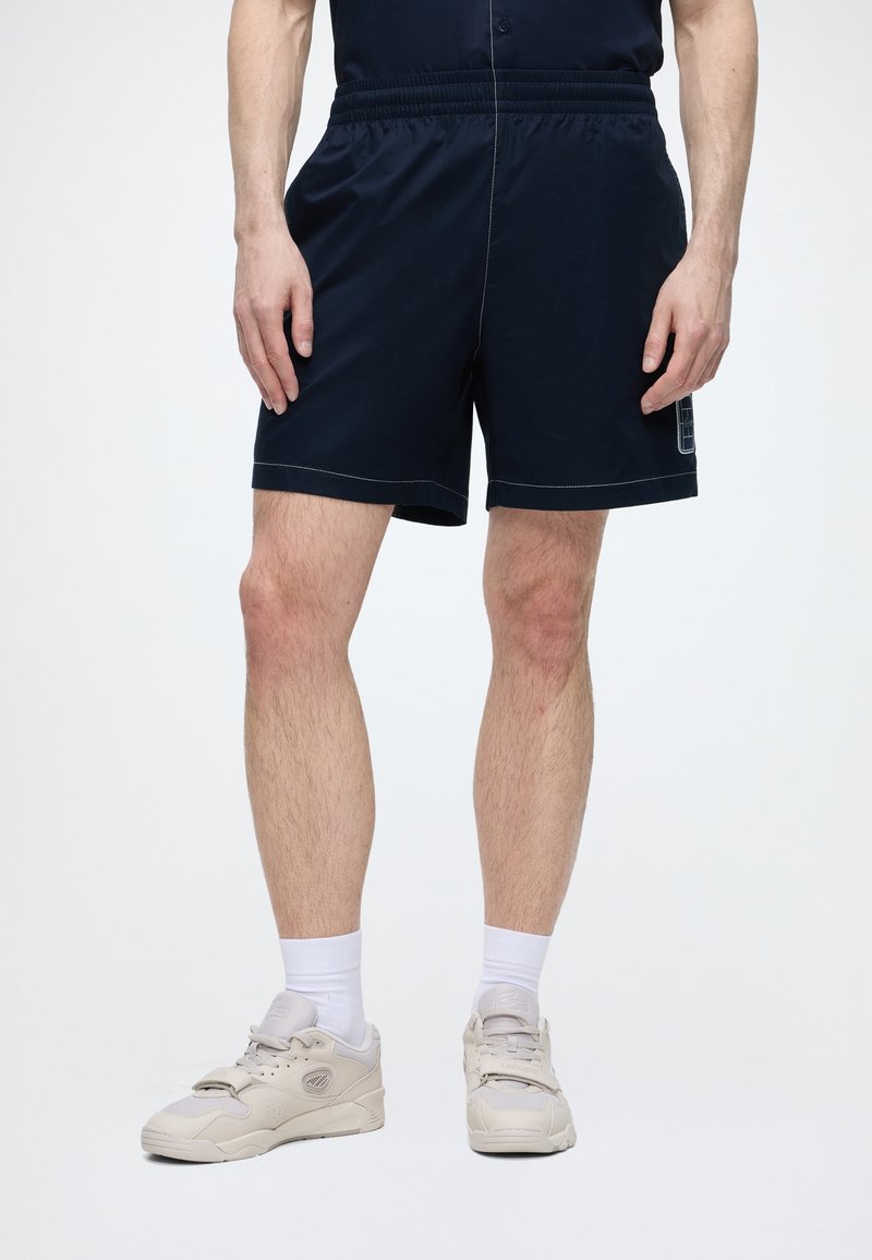 Homme portant un short noir, des chaussettes blanches et des baskets beiges, debout devant un fond clair uni.