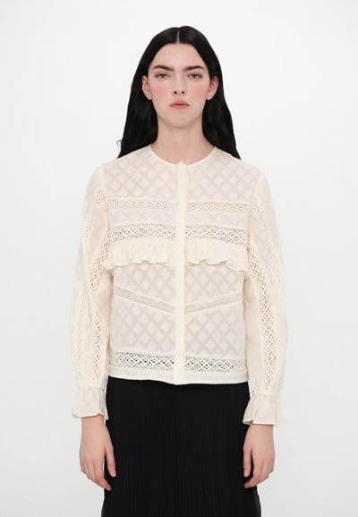 Blouse en dentelle crème avec un motif en diamant texturé, des accents volants et une fermeture à boutons. Manches longues avec des poignets délicats.