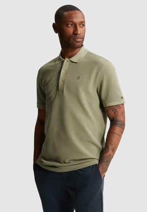 Muž s tetovaním, ktorý nosí svetlozelené textúrované polo s krátkym rukávom a tmavé nohavice, stojí s rukami v kapsách a pozerá sa doprava.