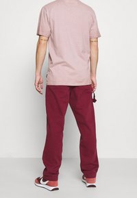 Herrenhemd in hellrosa mit kurzen Ärmeln, kastanienbraune Hose mit elastischem Bund und rot-weiße Sneakers. Einfaches Design ohne sichtbare Muster.