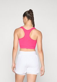 Kvinne med lang flette hestehale som har på seg en rosa racerback treningstopp og hvite sportshorts, snudd bort fra kamera mot en ensfarget bakgrunn.