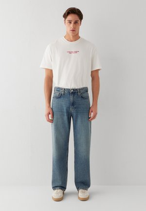 Junger Mann trägt ein weißes T-Shirt mit rotem Text und blaue gerade geschnittene Jeans, steht vor einer schlichten hellen Wand.