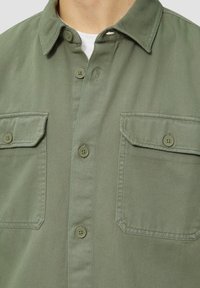 Olijfgroene geknoopte overhemd met een kraag, twee borstzakken en een gestructureerde stof. Voorzien van beige knopen en zichtbare stikseldetails.