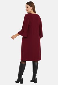 Robe bordeaux avec des manches évasées, longueur genoux, en tissu doux. Portée avec des bottes noires à hauteur de genoux. Design simple, sans motifs ni embellissements.
