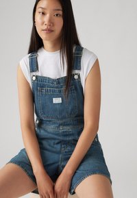 Denimski kombinezon v srednji modri barvi z nastavljivimi naramnicami, sprednjimi žepi in majhno nalepko. V kombinaciji s prilegajočo belo majico.