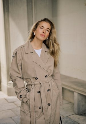 Ung kvinde med langt blondt hår iført en beige trenchcoat med hænderne i lommerne, stående i en urban stenkorridor.