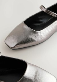 Metalické šedé baleríny se čtveratou špičkou, ozdobené tenkým páskem. Povrch má texturovaný finiš a lesklý vzhled.
