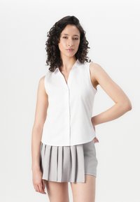 Camicia bianca senza maniche con colletto e chiusura con bottoni, abbinata a una gonna grigia plissettata. Tessuto morbido e design strutturato.