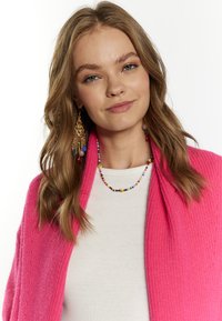 Ljusrosa, texturerad cardigan över en vit topp, med en flerfärgad pärlkrage och långa, intrikata guldörhängen. Släta hår.