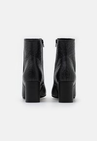 Botas negras hasta el tobillo con un acabado de cuero sintético texturizado. Tienen un tacón ancho y una cremallera en la parte trasera para facilitar el uso.