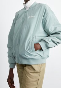 Veste bomber bleu clair avec fermeture éclair, poignets et taille côtelés, dotée de deux poches latérales et d'un logo sur la poitrine.