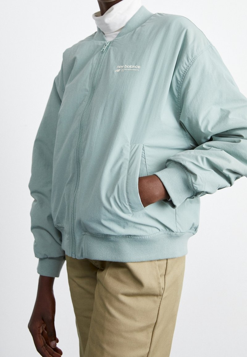 Veste bomber bleu clair avec fermeture éclair, poignets et taille côtelés, dotée de deux poches latérales et d'un logo sur la poitrine.
