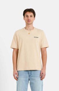 Camiseta de manga corta beige hecha de suave algodón, con un pequeño logo verde en el lado izquierdo del pecho y un corte holgado y cuadrado.