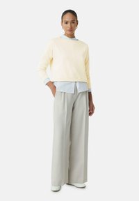 Afgeknotte gele sweatshirt met lange mouwen over een lichtblauw overhemd. Brede lichtgrijze broeken en witte schoenen maken de outfit compleet.