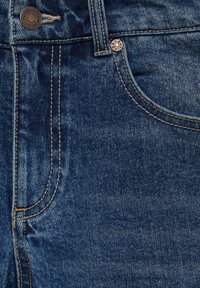 Los vaqueros de mezclilla azul oscuro presentan costuras visibles, un botón de color cobre y una textura ligeramente desvaída cerca del área del bolsillo frontal.