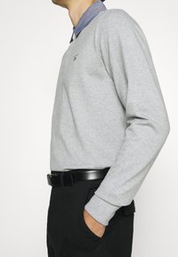 Harmaa neule pyöreällä kaula-aukolla, jossa on denim-paita. Kangas vaikuttaa pehmeältä, pitkät hihat ja ribbireunat. Musta vyö on käytössä.