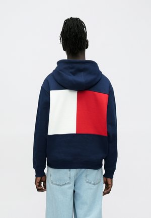 Άτομο με σκούρο δέρμα και dreadlocks φοράει μπλε σκούρο φούτερ με μεγάλο λευκό και κόκκινο μπλοκ στην πλάτη και ανοιχτό μπλε τζιν, με την πλάτη γυρισμένη.