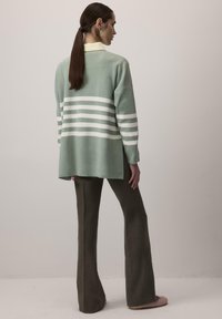 Muntgroene trui met witte horizontale strepen, relaxte fit, lange mouwen, gelaagd over een gele overhemd met kraag, gecombineerd met bruine flared broek.