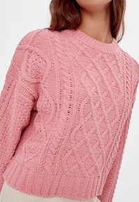 Suéter de punto trenzado rosa claro con escote redondo, patrones texturizados, hombros caídos y puños y dobladillo acanalados.