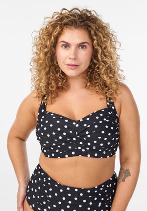 Krøllet kvinde med tatovering iført sort og hvidt bikini top med prikker og højtaljede underdele, stående mod en hvid baggrund.