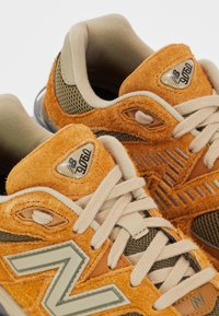 Gros plan sur des baskets New Balance 9060 orange et beige en suède et mesh avec des lacets beige clair.