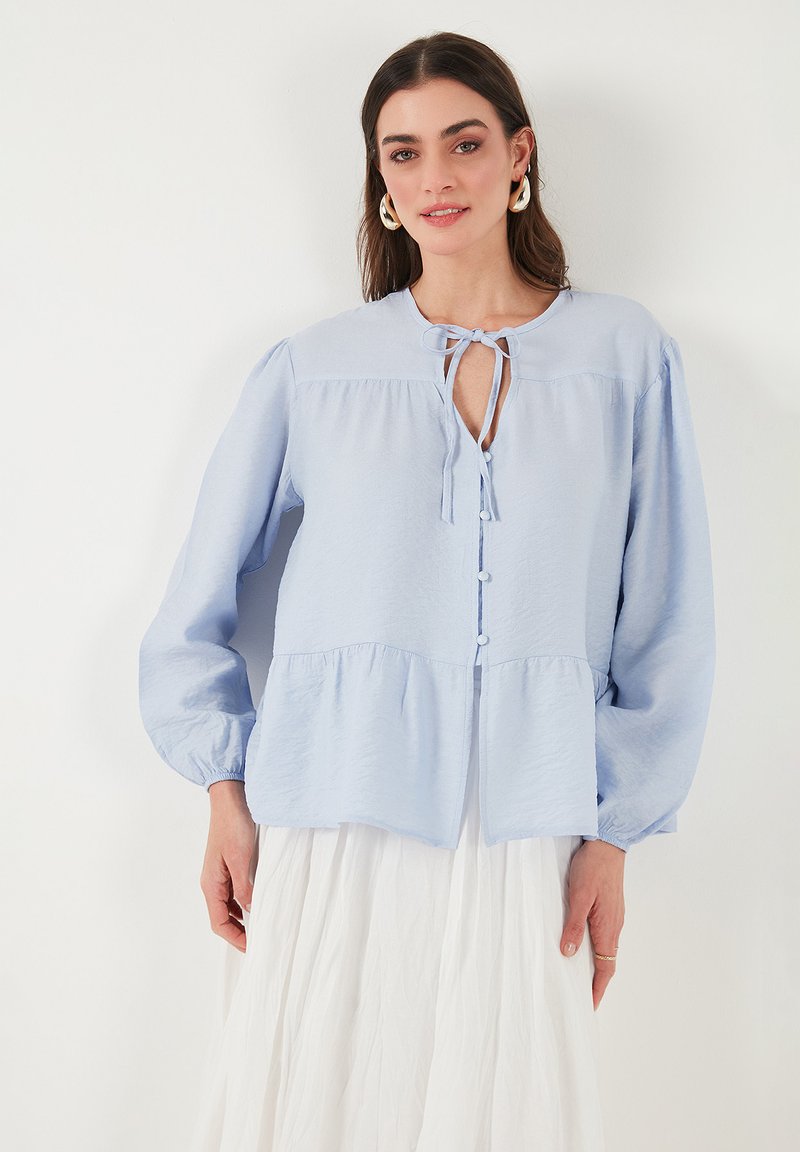 Blusa azzurro chiaro a maniche lunghe con collo a fiocco, frontale con bottoni e vita arricciata. Tessuto morbido e strutturato con vestibilità morbida.